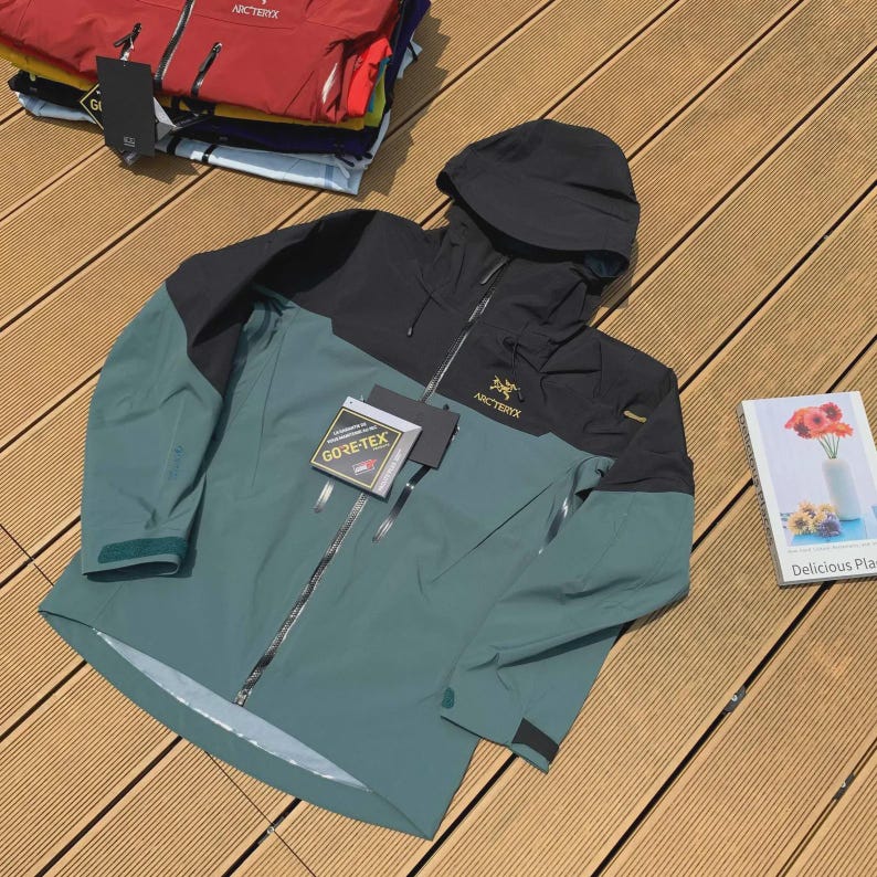 May include: A teal and black hooded jacket with a full-length zipper. The jacket has the brand name "Arc'teryx" on the chest and a "GORE-TEX" tag. Several other jackets are stacked in the background.