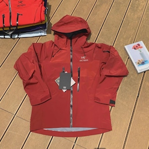 May include: A red hooded jacket with a black zipper and two front pockets. The jacket has the brand name "Arc'teryx" printed on the chest and hood. A tag is attached to the zipper. Several other jackets are stacked in the background.