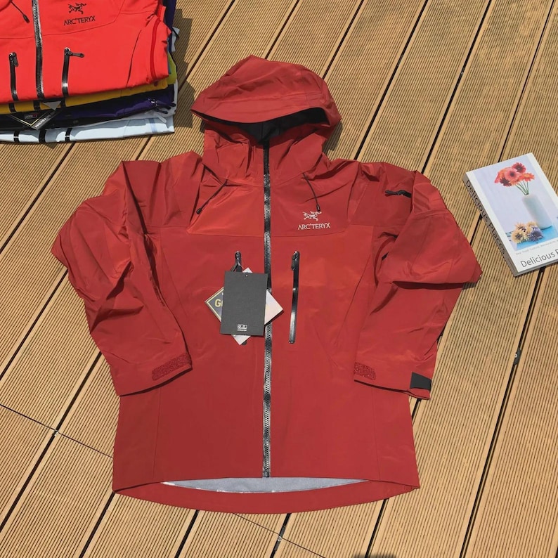 May include: A red hooded jacket with a black zipper and two front pockets. The jacket has the brand name "Arc'teryx" printed on the chest and hood. A tag is attached to the zipper. Several other jackets are stacked in the background.