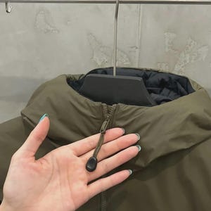 May include: An olive green jacket with a black lining, suspended from a metal hanger. The jacket features a high collar and a zip closure with a black toggle. The material appears smooth and possibly water-resistant.