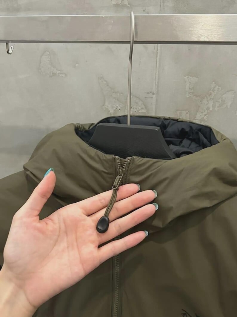 May include: An olive green jacket with a black lining, suspended from a metal hanger. The jacket features a high collar and a zip closure with a black toggle. The material appears smooth and possibly water-resistant.