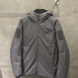 May include: A grey hooded jacket with a full-length zip, displaying the Arc'teryx logo on the chest. The jacket has two square patches on the sleeves and a small orange pull tab on the zip. The jacket is hanging on a wall.