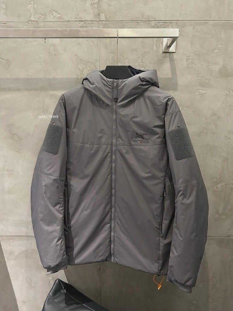 May include: A grey hooded jacket with a full-length zip, displaying the Arc'teryx logo on the chest. The jacket has two square patches on the sleeves and a small orange pull tab on the zip. The jacket is hanging on a wall.