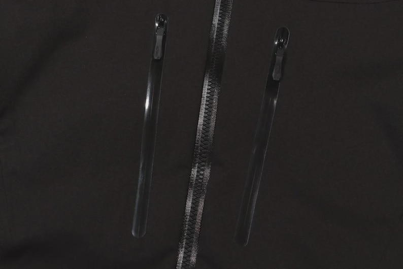 May include: Close-up of a black garment featuring a central zipper and two parallel, sleek, black zippers on either side. The zippers have metallic silver teeth and black pulls, set against a dark background.