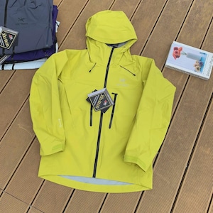 May include: A bright yellow hooded jacket with a black zipper and black accents. The jacket has a GORE-TEX tag attached. Additional jackets in shades of blue and purple are visible in the background.
