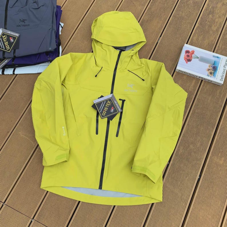 May include: A bright yellow hooded jacket with a black zipper and black accents. The jacket has a GORE-TEX tag attached. Additional jackets in shades of blue and purple are visible in the background.