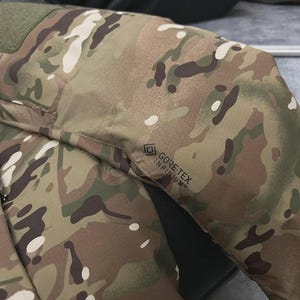 May include: Close-up of a camouflage jacket sleeve. The jacket features a pattern of brown, green, and white shapes. The GORE-TEX INFINIUM logo is visible on the sleeve. The jacket appears to be made of a durable, weather-resistant material.