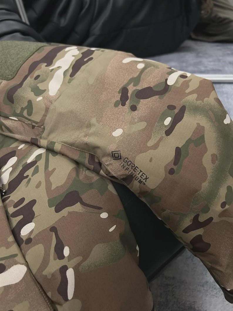 May include: Close-up of a camouflage jacket sleeve. The jacket features a pattern of brown, green, and white shapes. The GORE-TEX INFINIUM logo is visible on the sleeve. The jacket appears to be made of a durable, weather-resistant material.
