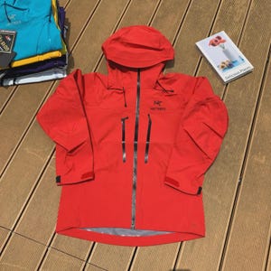 May include: A bright red hooded jacket with a full-length zipper and two vertical zippered pockets. The jacket has the brand name "Arc'teryx" printed on the chest. A stack of colorful jackets is visible in the background.