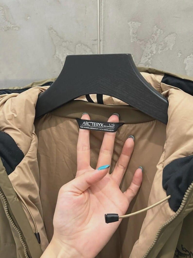 May include: An olive green and tan jacket with a black hood and a black zipper. The jacket is hanging on a black hanger. The tag reads "ARCTERYX" and "L/G".