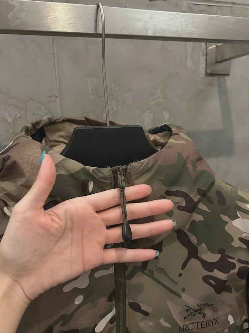 May include: Camouflage jacket in shades of green, brown, and tan, with a zip closure and high collar. The jacket displays the brand name "ARC'TERYX" on the lower right. The jacket is hanging on a black hanger.