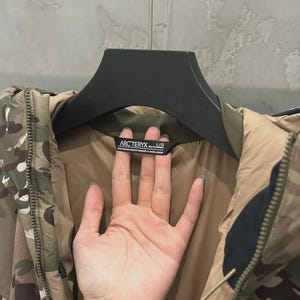 May include: Camouflage jacket with a brown lining and a black hanger. The jacket has a zip and the brand name "Arc'teryx" on a label. The jacket is a large size.