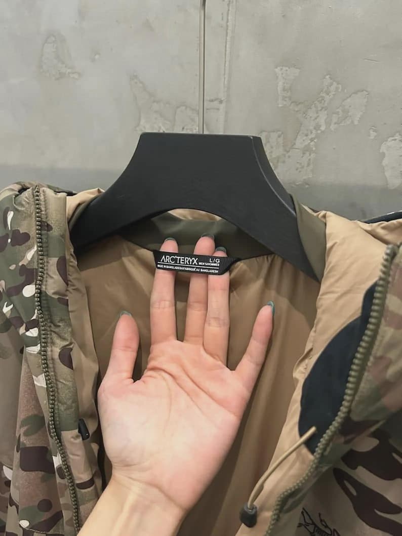 May include: Camouflage jacket with a brown lining and a black hanger. The jacket has a zip and the brand name "Arc'teryx" on a label. The jacket is a large size.