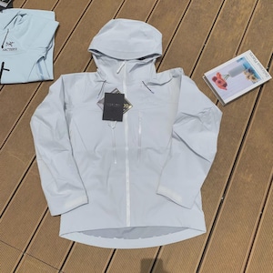 May include: A light blue hooded jacket with a full-length zipper, two side pockets, and a tag. The jacket is made of a lightweight material and is laid out on a wooden surface. A light blue shirt is visible in the background.