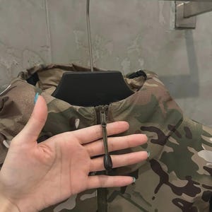 May include: Camouflage jacket in shades of green, brown, and tan, with a zip closure and high collar. The jacket displays the brand name "ARC'TERYX" on the lower right. The jacket is hanging on a black hanger.