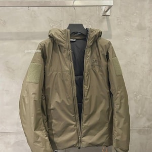 May include: A khaki-coloured hooded jacket with a full-length zip, patch pockets, and a small logo on the chest. The jacket is hanging on a metal hanger against a grey wall. The jacket has a black interior and a small orange tag.