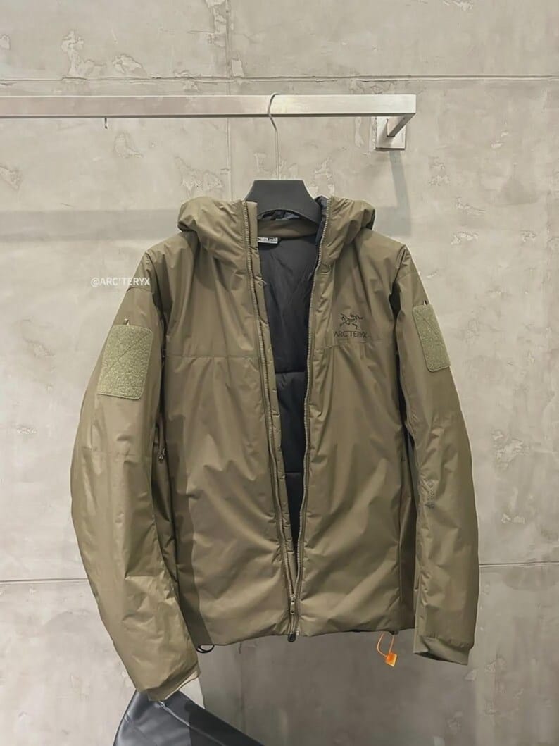 May include: A khaki-coloured hooded jacket with a full-length zip, patch pockets, and a small logo on the chest. The jacket is hanging on a metal hanger against a grey wall. The jacket has a black interior and a small orange tag.