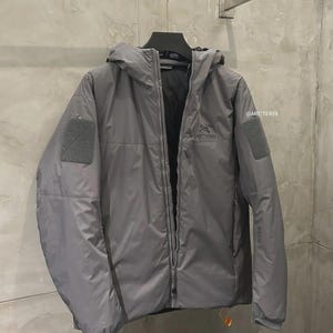 May include: A grey hooded jacket with a full-length zip and two square patches on the sleeves. The jacket has the brand name "Arc'teryx" printed on the chest and sleeve. The jacket is hanging on a metal rack.