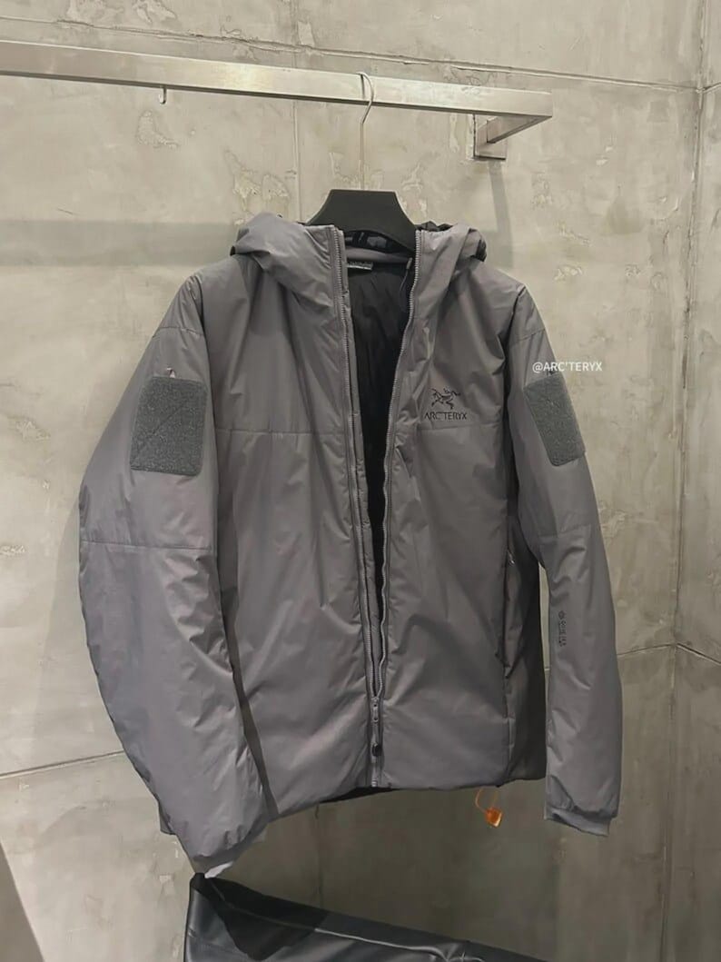 May include: A grey hooded jacket with a full-length zip and two square patches on the sleeves. The jacket has the brand name "Arc'teryx" printed on the chest and sleeve. The jacket is hanging on a metal rack.