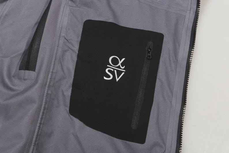May include: Close-up of the interior of a gray jacket. A black pocket with a zipper is visible, featuring the white letters "&alpha;SV". The jacket's interior is a light gray color.