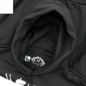 Pu&ograve; includere: Felpa con cappuccio nera con coulisse. La felpa presenta un emblema bianco con la lettera "M" al centro. Il cappuccio &egrave; aperto, rivelando l'interno del capo. La felpa &egrave; realizzata in un materiale morbido.