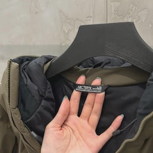 May include: Olive green hooded jacket with a black interior and a black zip. The jacket has a black label that says "ARCTERYX" and "L/G". The jacket is hanging on a black hanger.