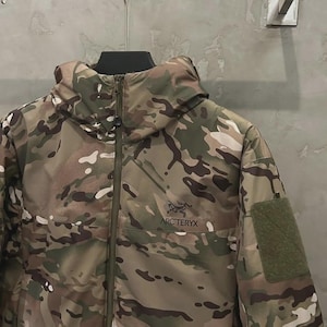 May include: A camouflage jacket with a hood and a zip fastening. The jacket has a green patch on the sleeve and the "ARC'TERYX" logo. It is hanging on a metal hanger against a grey wall.