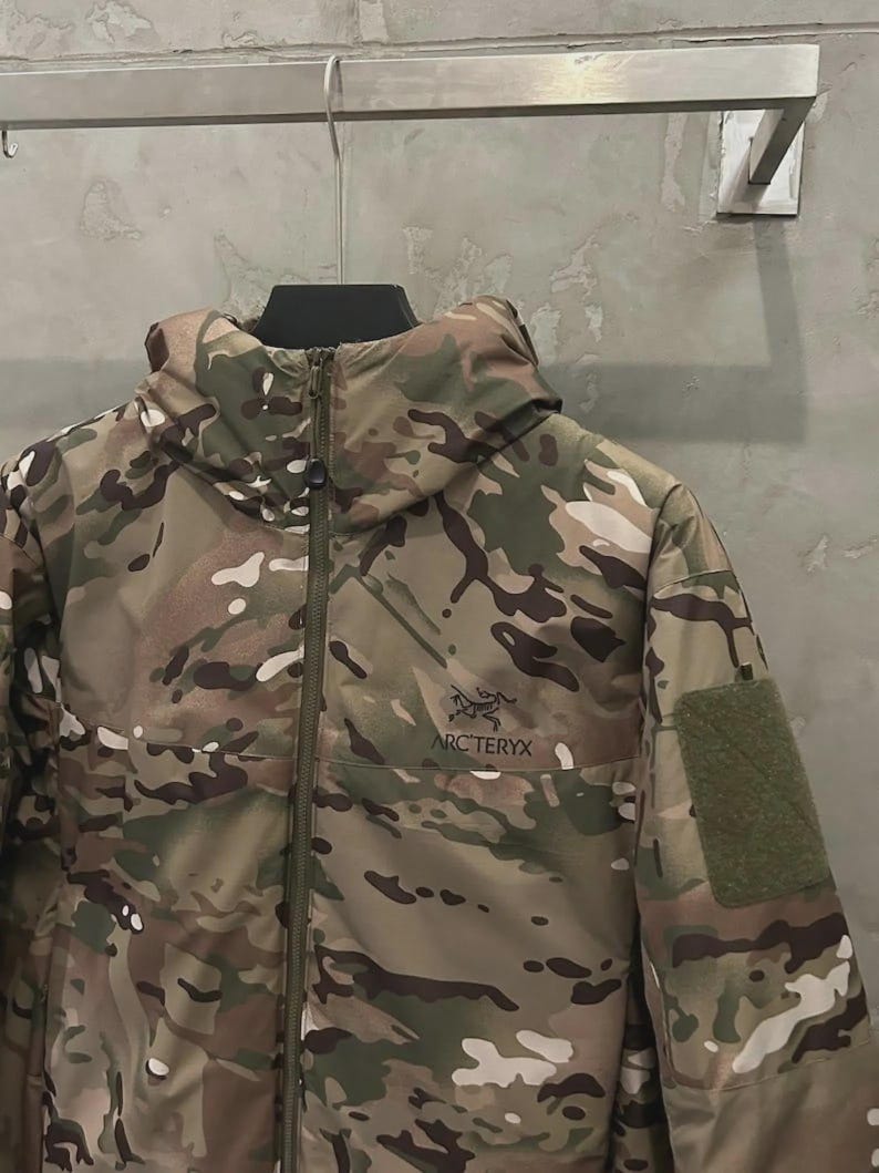 May include: A camouflage jacket with a hood and a zip fastening. The jacket has a green patch on the sleeve and the "ARC'TERYX" logo. It is hanging on a metal hanger against a grey wall.