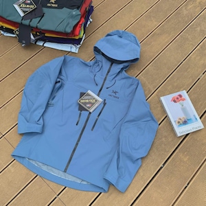 May include: A light blue Arc'teryx Gore-Tex jacket with a hood and a black zipper. The jacket has two front pockets and the Arc'teryx logo on the chest. A stack of other jackets is visible in the background.