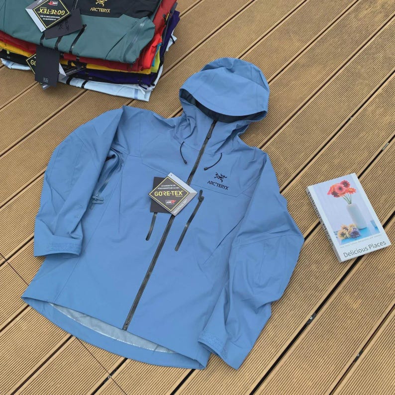 May include: A light blue Arc'teryx Gore-Tex jacket with a hood and a black zipper. The jacket has two front pockets and the Arc'teryx logo on the chest. A stack of other jackets is visible in the background.