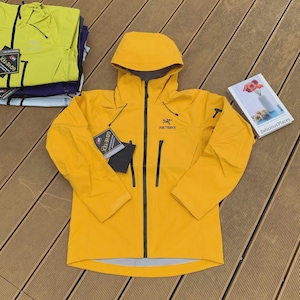 May include: A bright yellow hooded jacket with a black zipper and pockets. The jacket has the brand name "Arc'teryx" on the chest and a "GORE-TEX" tag. A book titled "Delicious Places" is also visible.