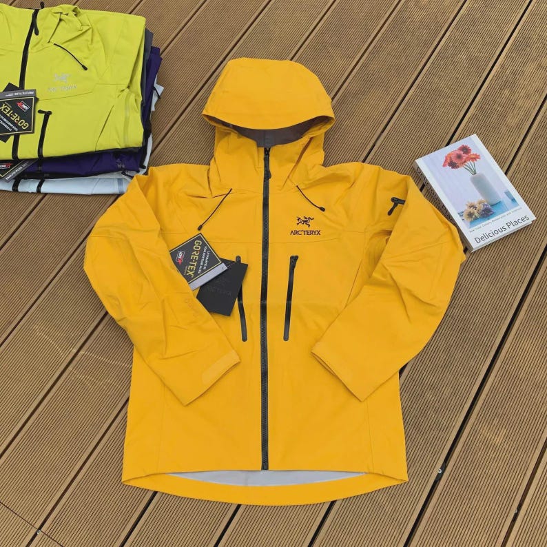May include: A bright yellow hooded jacket with a black zipper and pockets. The jacket has the brand name "Arc'teryx" on the chest and a "GORE-TEX" tag. A book titled "Delicious Places" is also visible.