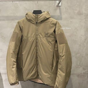 May include: A tan jacket with a hood and a zip fastening. The jacket features a logo on the chest and patches on the sleeves. The jacket is displayed on a hanger. The brand is Arc'teryx.