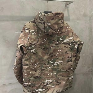 May include: A camouflage jacket with a hood, in shades of brown, green, and white, hangs on a metal rail. The jacket has a full-length zip and a high collar. The @ARCTERYX logo is visible on the left side of the jacket.