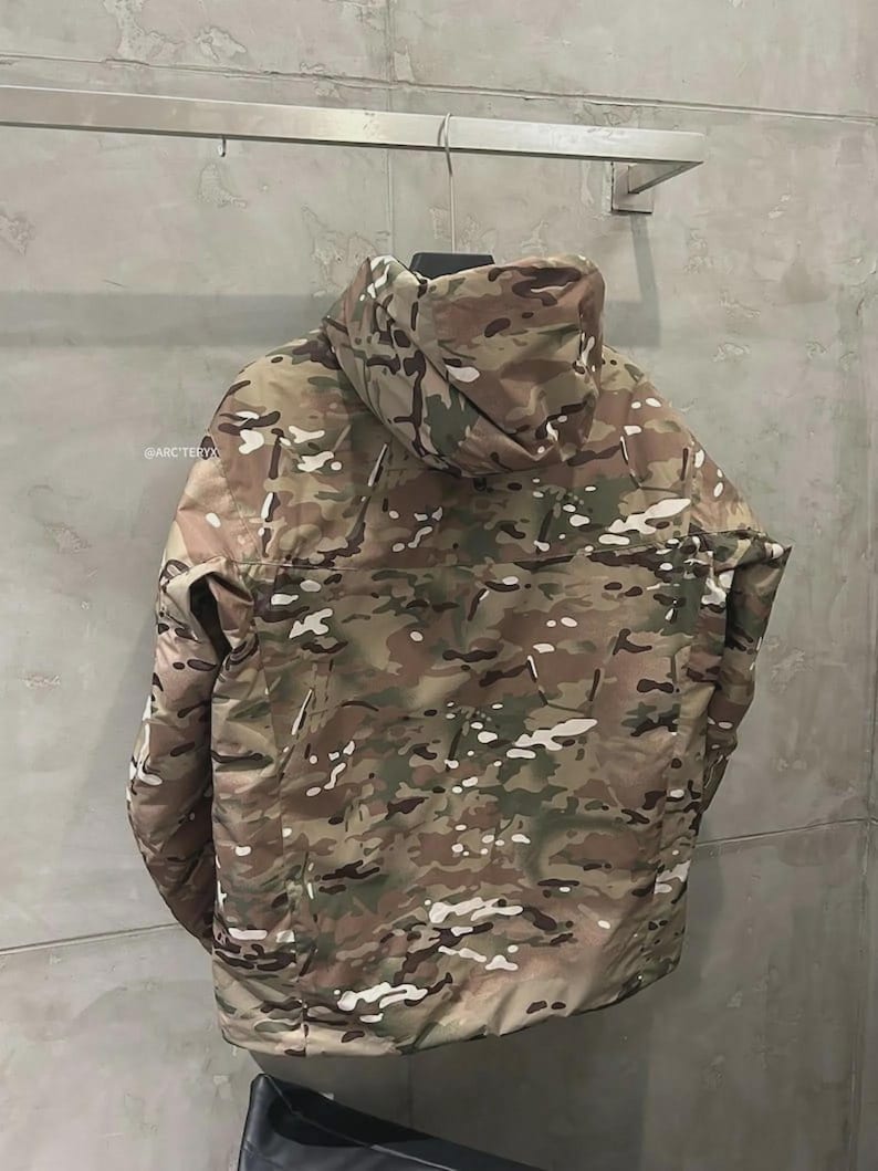 May include: A camouflage jacket with a hood, in shades of brown, green, and white, hangs on a metal rail. The jacket has a full-length zip and a high collar. The @ARCTERYX logo is visible on the left side of the jacket.