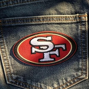 May include: A dark blue denim pocket featuring an embroidered oval patch. The patch displays the letters SF in white with a black outline, set against a red background, all enclosed within a black and red border. The denim fabric has visible stitching.