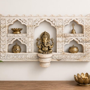 May include: A rectangular, light-coloured wooden wall shelf featuring carved arched niches. A brass Ganesha statue sits in the centre. The shelves hold a brass lotus-shaped bowl, a small brass bell, and two ornate brass lidded containers.
