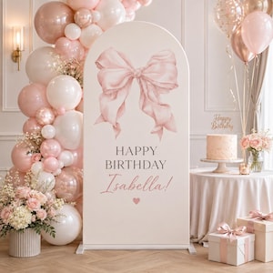 Op de afbeelding: Een boogvormige achtergrond met een grote roze strik in aquarel-stijl, de tekst "HAPPY BIRTHDAY Isabella!" en een klein hartje. Het geheel is omringd door een tros roze, witte en metallic roségouden ballonnen en een bloemstuk.