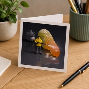 Puede incluir: Una tarjeta de felicitaci&oacute;n cuadrada que muestra una figura en miniatura de un trabajador con chaqueta amarilla y pantalones grises usando una hidrolavadora para limpiar una golosina de goma naranja y amarilla azucarada sobre una superficie oscura.