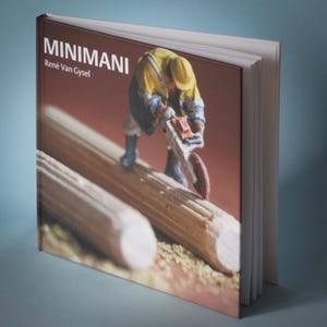 Puede incluir: Un libro de tapa dura titulado "MINIMANI" de René Van Gysel. La portada presenta una figura en miniatura con una chaqueta amarilla y botas azules usando una motosierra sobre una clavija de madera. La portada del libro es de color marrón rojizo.