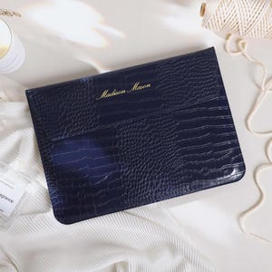May include: A dark navy blue rectangular laptop sleeve with a textured crocodile skin pattern. The front flap features the gold embossed name Madison Mason. The sleeve is positioned on a white fabric surface alongside a candle and string.