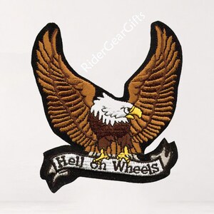 May include: An embroidered patch featuring a bald eagle with brown wings spread wide, a white head, and a yellow beak. Below the eagle is a white banner with black borders and the text "Hell on Wheels" written in a bold, black, stylized font.