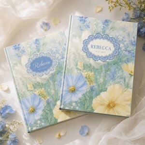 Könnte beinhalten: Zwei Notizbücher mit festem Einband und einem Aquarell-Blumenmuster in Blau-, Gelb- und Grüntönen. Jeder Einband zeigt einen zentralen, verzierten blauen Rahmen mit dem Namen REBECCA und dem Text EST 2024 in einer klaren Serifenschrift.