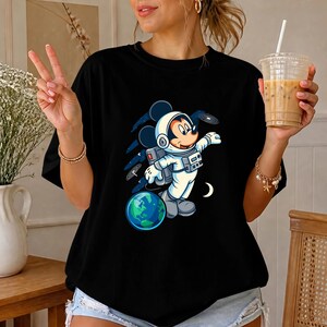 May include: A black t-shirt featuring a graphic print of a cartoon mouse wearing a white astronaut suit, floating in space near a blue and green planet. The design includes dark blue star trails and a small white crescent moon against the black fabric.