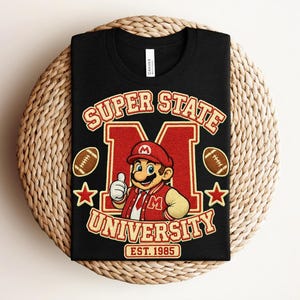 May include: A black t-shirt featuring a graphic of a cartoon character wearing a red cap and varsity jacket, centered in front of a large letter M. The text reads Super State University Est. 1985, accompanied by two footballs and two stars.