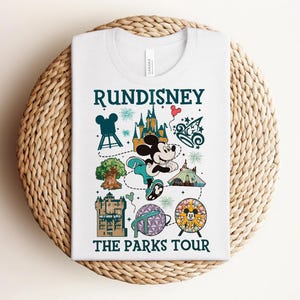 May include: A white t-shirt featuring a graphic print with the text RUNDISNEY THE PARKS TOUR. The design includes a central illustration of Mickey Mouse running, surrounded by various park icons, a castle, a tree, and fireworks in teal and tan.