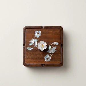 May include: A square dark brown wooden ashtray featuring an inlaid mother-of-pearl floral design. The inlay consists of two flower stems with leaves and a central flower blossom, all displaying iridescent white and blue color tones.
