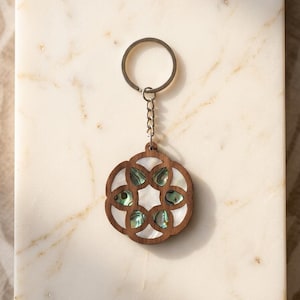 May include: A circular keyring featuring a dark brown wooden frame with a geometric cutout design. The centre is inlaid with iridescent abalone shell pieces and white mother-of-pearl accents. It includes a silver-toned metal split ring and chain.