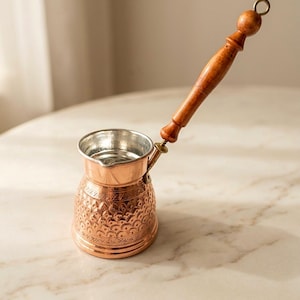 May include: A copper coffee pot with a silver-toned interior and a long, turned wooden handle. The exterior of the pot features an intricate, embossed pattern of repeating semi-circular motifs and geometric lines around the base and body.