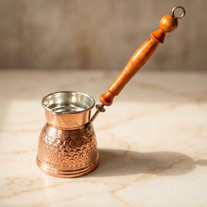 May include: A copper coffee pot with a silver-toned interior and a long, turned wooden handle. The exterior of the pot features a decorative embossed pattern of repeating semi-circles and geometric lines around the base and body.